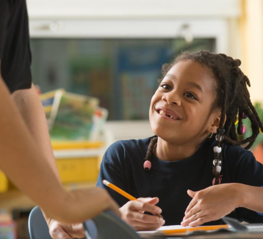 A girl sitting at a school desk looks up smiling at a teacher.